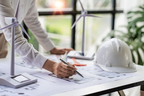 Young Asian architect with blueprints working on lapotp in office  Career working hard on wooden table, painting with windmills, designing model of renewable wind house building  and sunlight