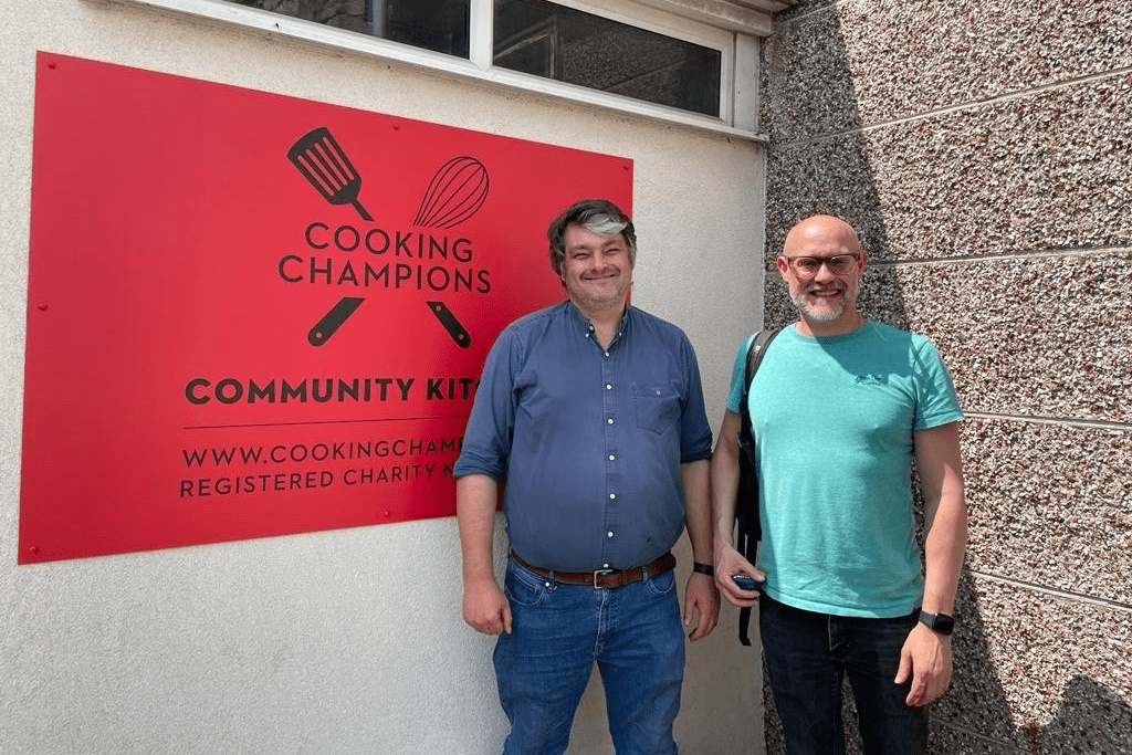 Peter Wiggins, wearing a blue shirt and blue denim jeans, and Rodney Unsworth, wearing a turquoise t-shirt and black trousers, stand in front of the red sign for Cooking Champions' community kitchen